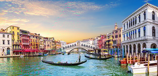 Gondolas glide through the Grand Canal in Venice, Italy, with the colorful facades of historic buildings and the Rialto Bridge in the background at sunset.
