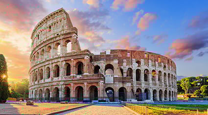 The Colosseum in Rome, Italy, glows in the golden light of dusk, highlighting its ancient arches and stone structure against a dramatic sky.
