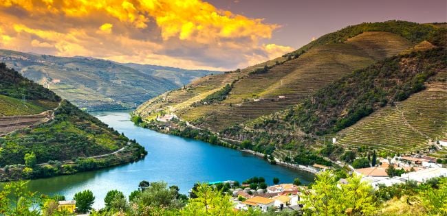 Scenic view of Portugal’s Douro Valley with a winding river, terraced vineyards, and hillside villages under a vibrant sunset sky.