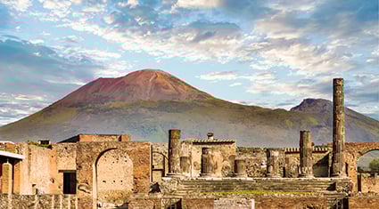 The ancient ruins of Pompeii with Mount Vesuvius in the background, captured under a partly cloudy sky.