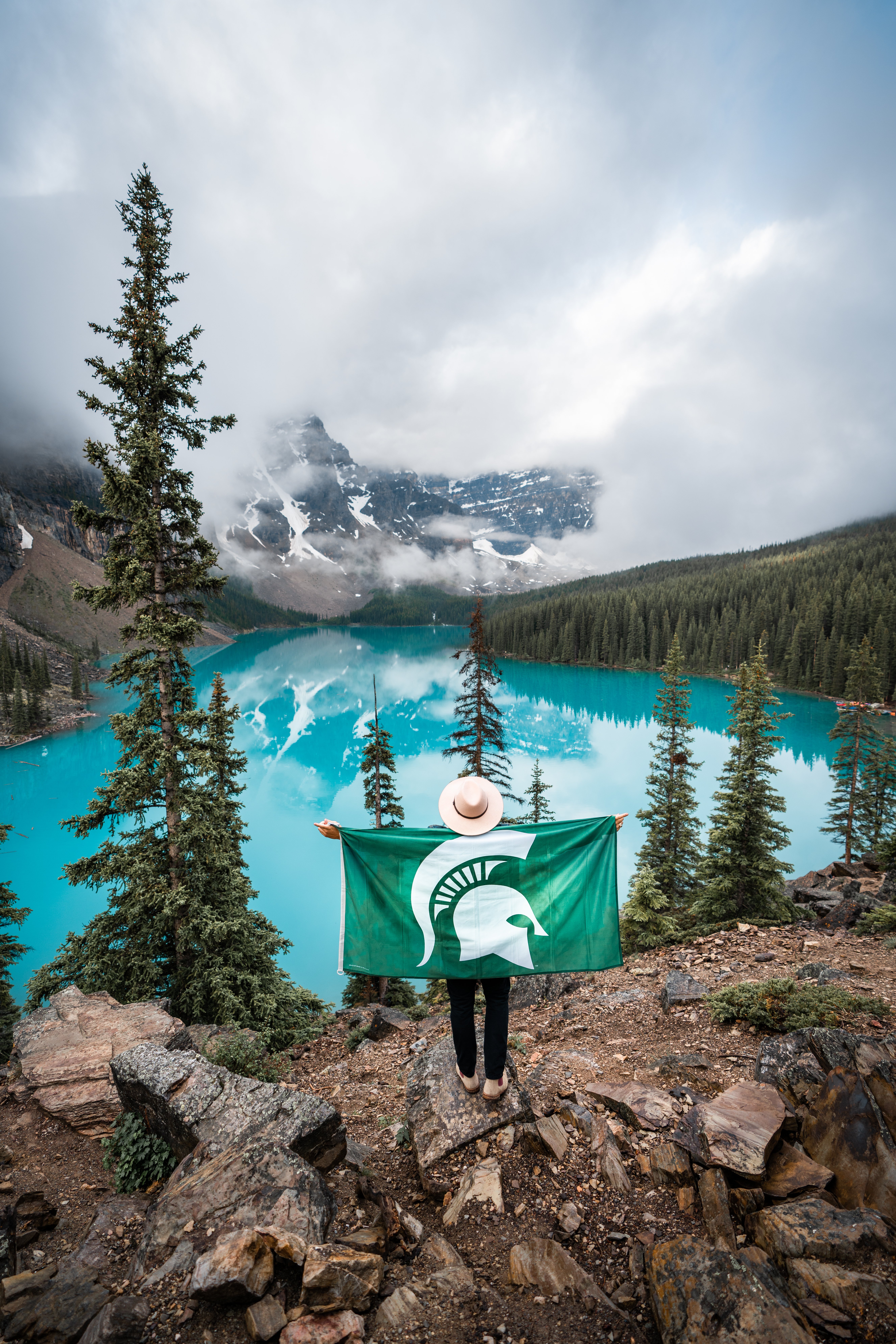 A person stands on a rocky overlook holding a Michigan State University flag, facing a stunning turquoise lake surrounded by evergreen trees and snow-capped mountains.