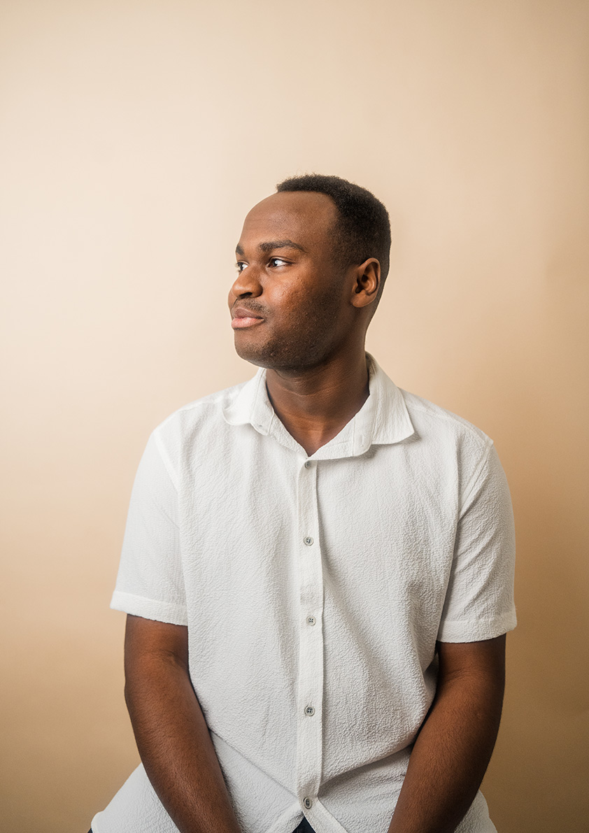 Christian Nderu sits in profile wearing white linen button-up looking left.