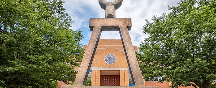 Close up of the Beta statue behind the college of engineering. In the middle of the of the statue is the engineering building. It is a sunny day with trees in front of the building