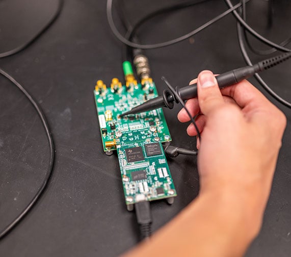 A student working on a circuit board, their hand is holding a tool to make edits to it