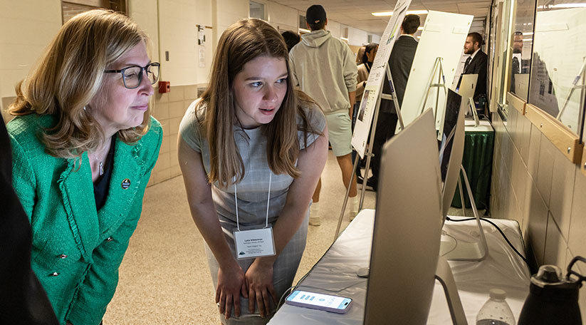 Interim President, Teresa Woodruff is looking at a computer screen with a engineering student. The student is sharing her capstone project during Spring 2023 Design Day