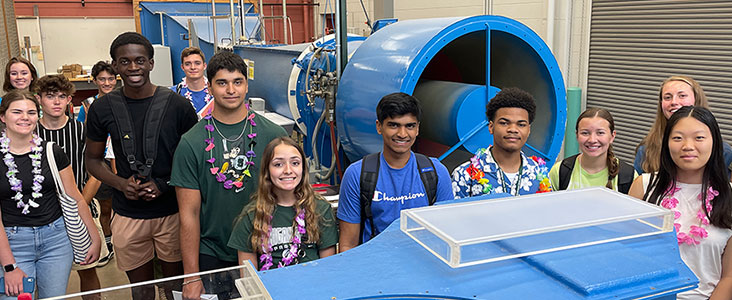 a wide range of multi-cultural students standing in a lab in front and behind two blue machines during a ESSA event