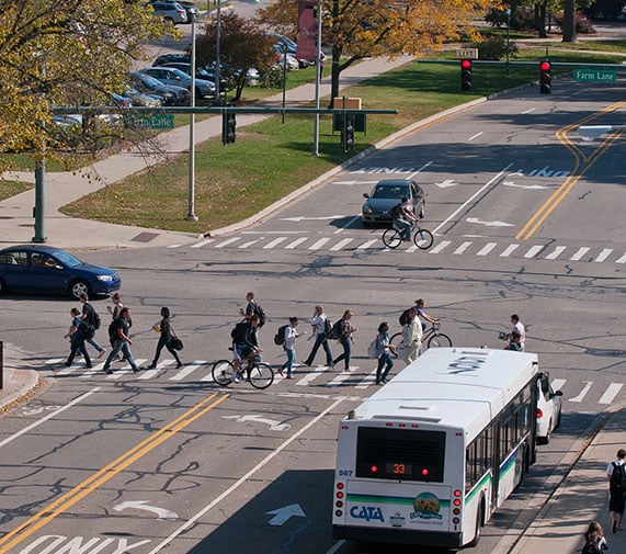birds eye view of the intersection of Farm lane and Shaw lane, MSU student are walking across the crosswalk. There is a CATA bus at a stop in front of the crosswalk. 
