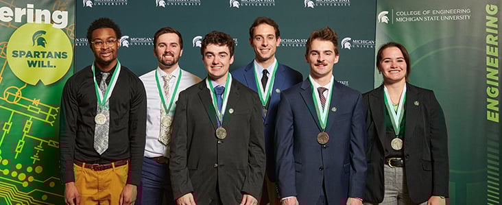 Award wining students at the Spring 2023 Design Day, there is 1 black male, 4 white male and 1 white female student standing behind a backdrop wearing their medals