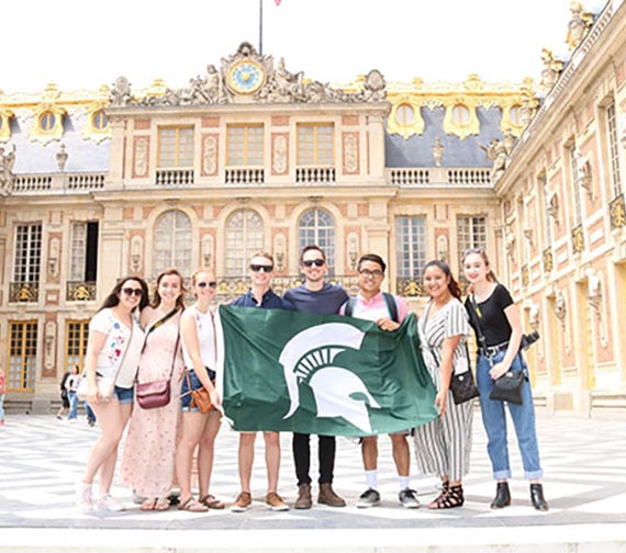 MSU Engineering student group holding a flag with the spartan helmet on it, standing in front of a building at CEA Paris Center