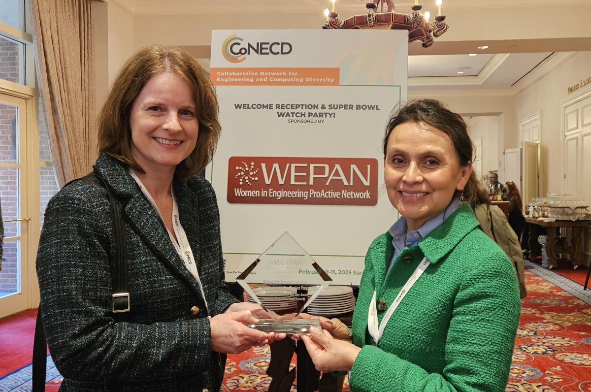 Two individuals hold an award between them and smile at the camera in front of a poster that reads 'WEPAN Women in Engineering ProActive Network.'
