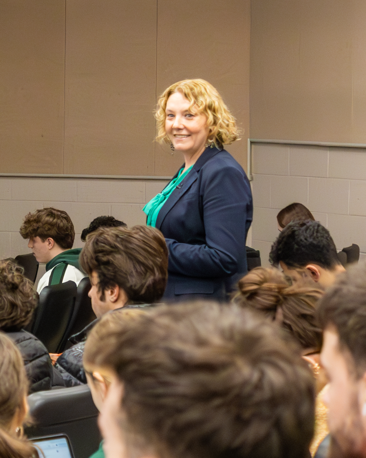 Professor with blonde hair wearing a blue blazer and green shirt standing in a classroom full of seated students