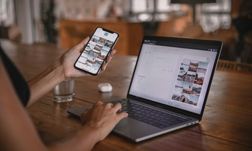 A user is working on a laptop while holding a smartphone, both devices showing multiple image thumbnails.