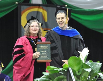 Todd Elder in doctorate robes receives a plaque symbolizing his award. 