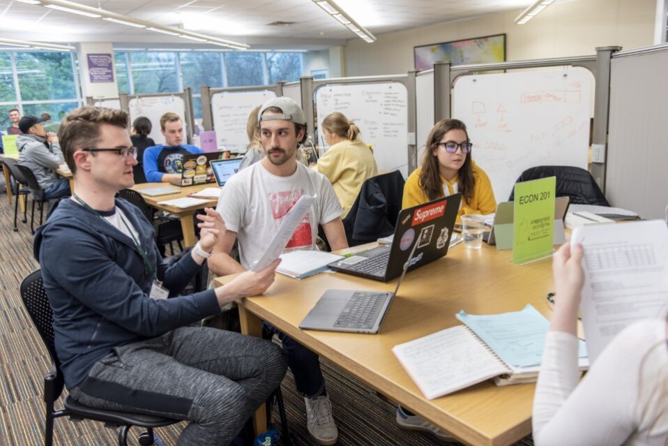 Two male students and a female student discuss their papers and another female student looks at her computer screen at an Econ 101 table. 