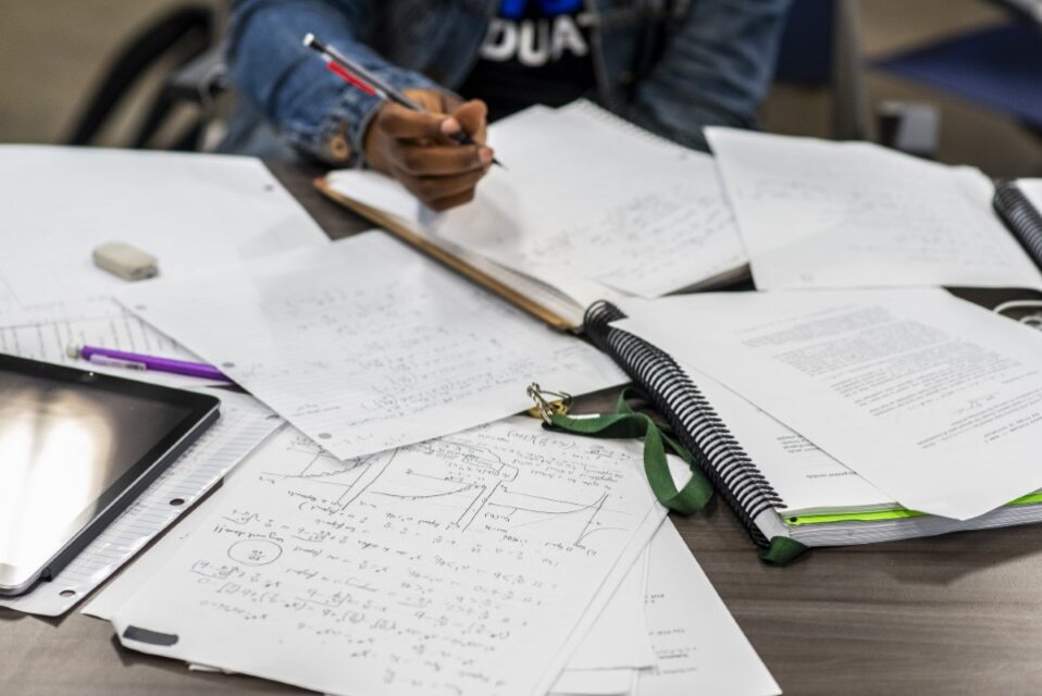 A view of a table with an Ipad, notebooks and book with the hands of a black female student writing. 