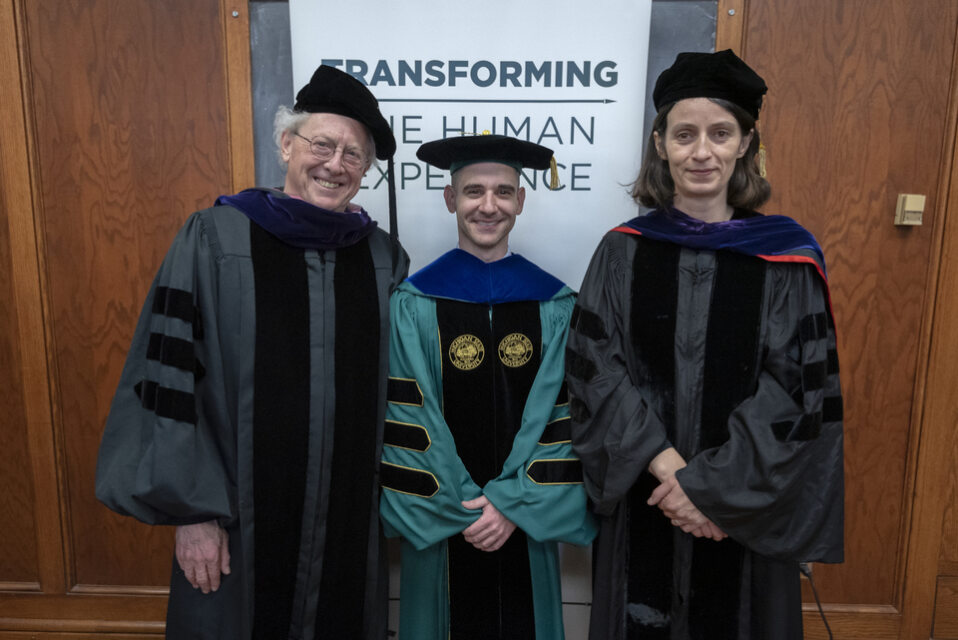 One white professor and two white PhD students in graduation robes, one man, one woman. 