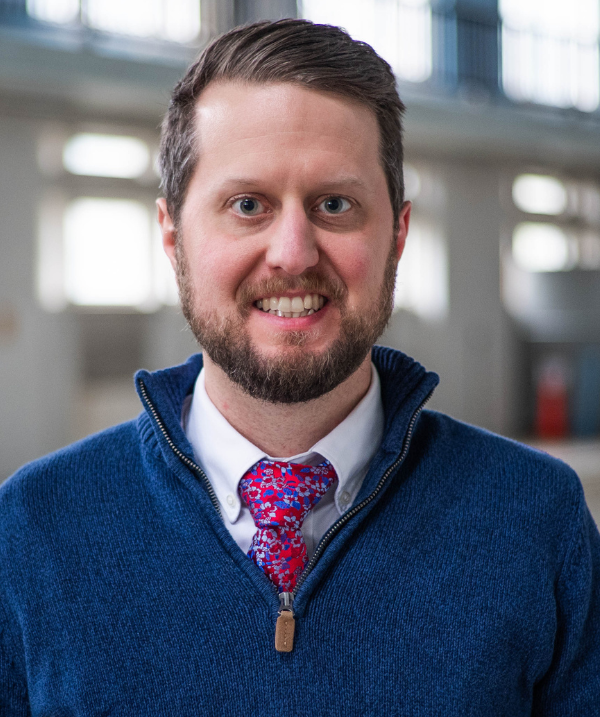 Matt Harkey headshot. Harkey stands in a gym and smiles toward the camera. Harkey wears a dark blue sweater over a white collared shirt.