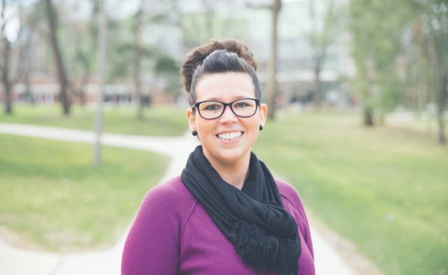 A smiling individual wearing glasses, a purple sweater, and a black scarf, standing outdoors with a paved walkway and trees in the background.