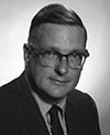 Black-and-white photo of a man in a suit and tie, wearing glasses and looking directly at the camera.