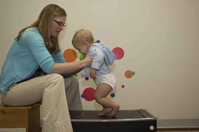 A young child learning to walk with the support of a researcher in a lab.