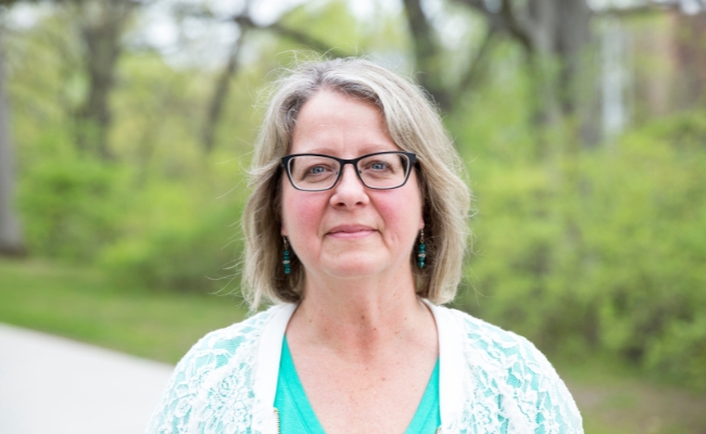 A smiling individual with shoulder-length light hair, wearing glasses, a turquoise top, and a white lace cardigan. They are standing outdoors with a blurred background of greenery and trees.