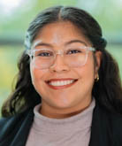 Yazmin Castruita Rios headshot. Yazmin stands in front of a window. Yazmin wears a beige turtleneck and clear-rimmed glasses. Yazmin's dark hair is long and falls past the shoulders.