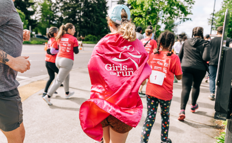 Picture of runners in a race. Girl in forefront of photo wears a pink cape with "Girls on the Run" logo.