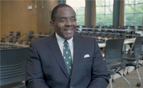 Billy Hawkins headshot. Billy wears a dark gray blazer over a white button-down shirt and an MSU green neck tie. Billy is seated in the Kiva, a circular, tiered classroom within Erickson Hall, home to the MSU College of Education.