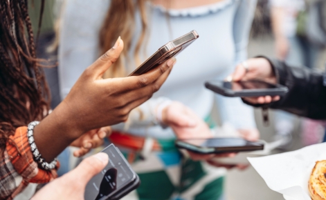 A group of people holding smartphones, engaging in an activity or conversation. The image focuses on their hands and devices, suggesting a shared interaction or moment involving mobile technology. The background is blurred, with some clothing details visible.
