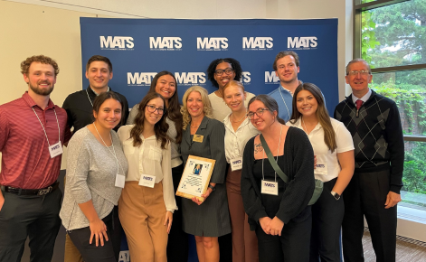 A group of smiling people posing for a photo indoors in front of a blue backdrop with the MATS logo repeated across it. In the center, a woman holds a plaque, surrounded by men and women wearing name tags. The setting includes a large window with a view of greenery outside.