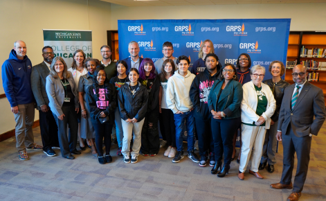e image features a group of individuals gathered together for a group photo. They are standing in front of a backdrop displaying the logos of Michigan State University (MSU) College of Education and Grand Rapids Public Schools (GRPS). The group includes both adults and students, with everyone smiling and posing for the camera. The adults appear to include faculty, staff, and school administrators, while the students represent the participants of the inaugural Community Teachers cohort program launching in October 2024.The initiative aims to accelerate the career paths of future educators, providing them with opportunities to gain experience and ultimately secure full-time teaching positions within the GRPS K-12 school system. The backdrop and surrounding setting suggest the photo was taken in a library or academic environment.