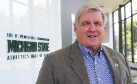 A middle-aged Caucasian man with short gray hair, smiling, stands in a corridor of the Michigan State Athletics Hall of Fame. He wears a light gray jacket over a blue checked shirt. The background features large windows looking out to an indoor area with green and white banners.