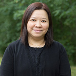 Gloria Lee headshot. Gloria smiles toward the camera in an outside photo. Gloria wears a black blouse with a necklace. Gloria's dark hair is about shoulder-length.