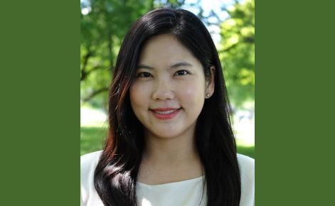 A portrait of a woman with long dark hair, smiling outdoors with green trees and sunlight in the background. She is wearing a light-colored blouse and has a friendly expression.