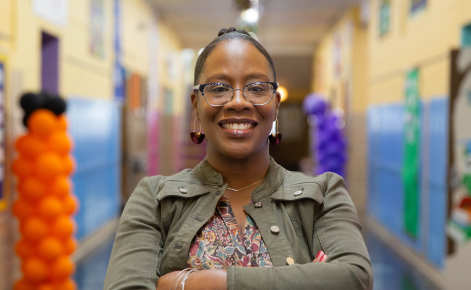 Candice Nicholson headshot. Candice wears a multicolored floral blouse under a dark green jacket with large buttons. Candice wears wire-rimmed glasses and large hooped earrings. Candice is standing in a school hallway.