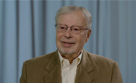 Rick Stiggins headshot. Stiggins wears a brown blazer over a white button-down shirt. Stiggins wears wire-rimmed glasses. Stiggins is seated against a blue curtain backdrop.