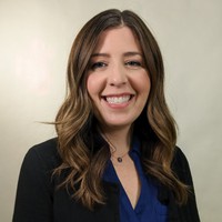 Dietzel headshot; Kathy is wearing a blue blouse and a black blazer. Her brown hair is wavy and falls below her shoulders. She stands agains a neutral background. 