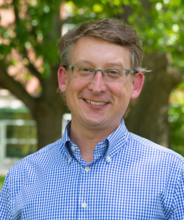 Cary Roseth headshot. Roseth stands outside in front of a tree with full foliage. Roseth wears a blue and white checkered button down shirt and glasses.