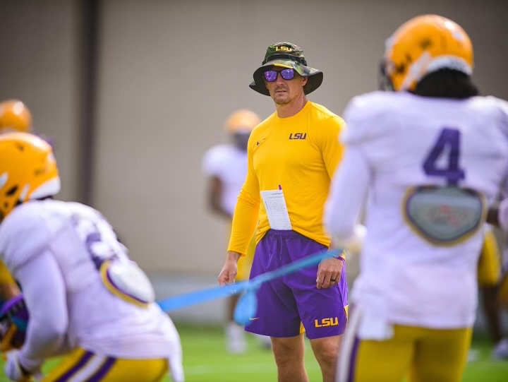 Bryson Peddy, wearing a bright yellow shirt and purple shorts, watches football players during practice.