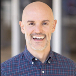 This is a headshot of Bill Marsland. Bill is bald, has light skin, and a short grey beard. He is wearing a dark blue and red plaid button down shirt. The background is blurry with 2 vertical white posts.