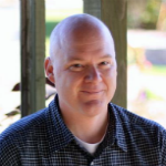 This is a headshot of Ron Houtman. He has light skin and is bald. He is looking at the camera and smiling. He is wearing a dark blue button down shirt. The background is foliage that is out of focus.