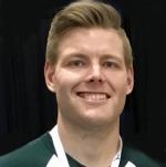 This is a picture of Kyle Shack. Kyle is smiling and looking at the camera. Kyle has blonde hair and light skin. He is wearing a green and white (MSU) shirt and in front of a dark background.