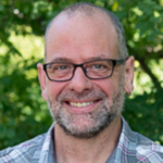 This is a headshot of Matthew Koehler. He has short grey hair, a short grey beard and light skin. Matt wears glasses an is smiling at the camera. He is wearing a blue and white button down shirt. He is outside with green foliage behind him.