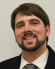 This is a picture of Michael Lachney. He has short brown hair, a short brown beard, and light skin. He is smiling and facing the camera. He is wearing a dark suit jacket, white shirt, and tie. The background is plain white.