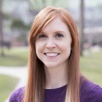 This is a headshot of Liz Boltz. She has long red hair and light skin. She is looking at the camera and smiling. She is wearing a purple shirt. The background is foliage that is out of focus.