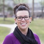 This is a headshot of Candace Robertson. She has dark hair that is up in a bun and light skin. She wears glasses. She is smiling and looking at the camera. She is wearing a purple sweater and black scarf. The background is foliage that is out of focus.