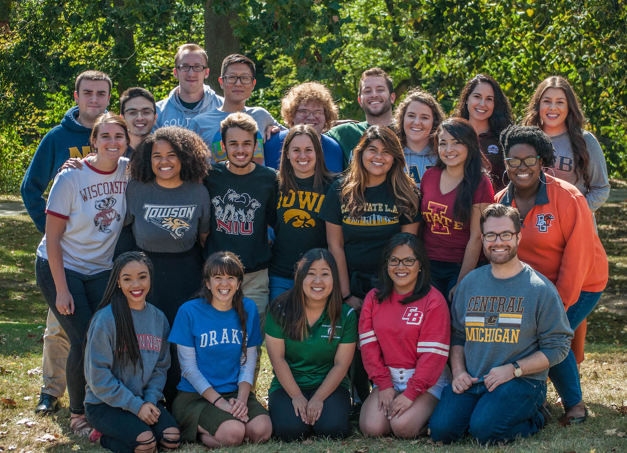 Group of students wearing various higher education-affiliated shirts pose for a group photo.