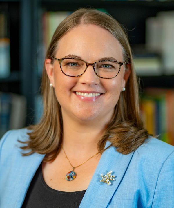 Madeline Mavrogordato headshot. Madeline wears a light blue blazer over a black blouse, as well as multicolored broach and necklace. Madeline wears dark-rimmed glasses. Madeline's blonde hair is about shoulder length. Madeline stands in front of a full bookshelf and a wall of decorative green tiles.