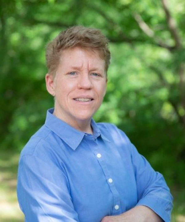 Kristen Renn headshot. Kristen wears a blue button down shirt. Kristen is standing outside and is smiling toward the camera.
