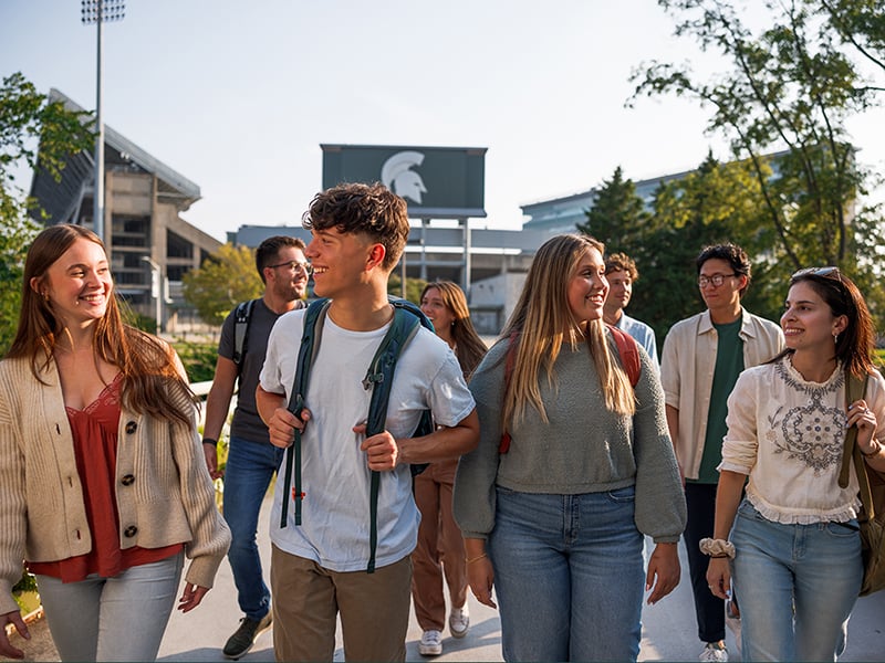 Group of students walking across bridge with Spartan helmet scoreboard in background